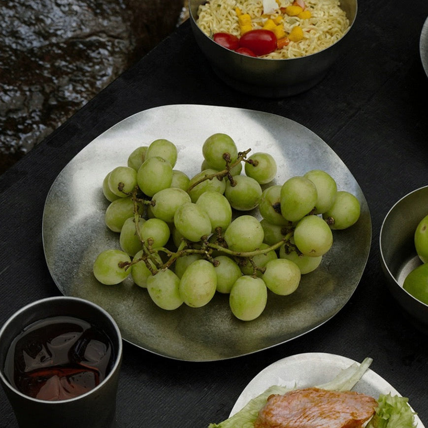 Bowl of salad with tomatoes, plate of green grapes, and other food items on a dark surface.