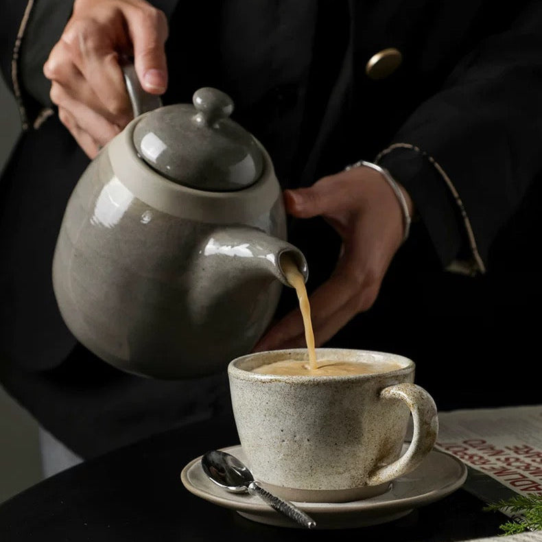 Person pouring tea from a gray ceramic teapot into a white mug on a table with a newspaper.